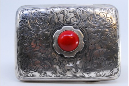 Cigarreira em prata com gravados vegetalistas, cabochão vermelho, de formato curvo e tampa basculante.