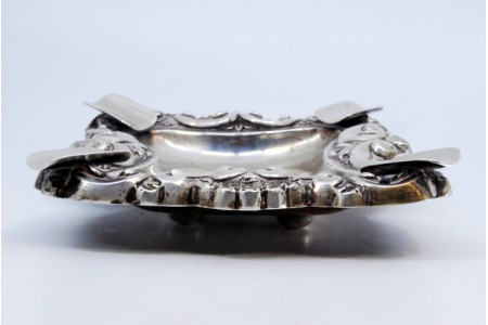 Cinzeiro em prata com relevos e aplicações, com quatro suportes para cigarros, e assente em quatro pés redondos.