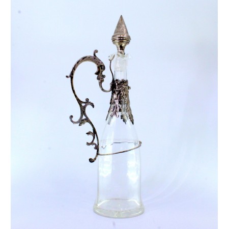 Garrafa de vinho em vidro lapidado com estrutura em prata e pega recortada em forma de figura mítica alada.