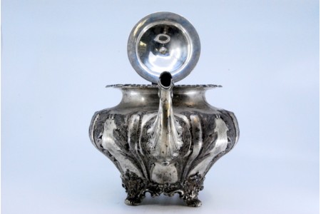 Bule em prata com aplicações na tampa e na pega, com quatro pés na base e relevos.