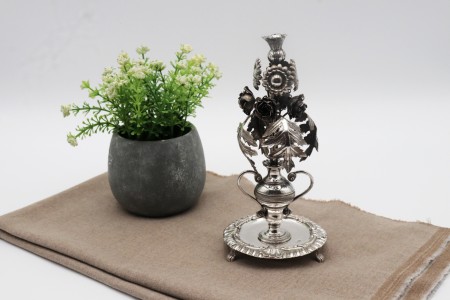 Paliteiro em prata com acanteiro de vulto perfeito, flores e folhas sobre jarra com duas asas e frisos.