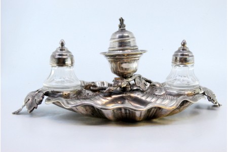 Tinteiro em prata com dois depósitos em vidro lapidado e campainha, cartela para monograma e assente sobre três pés em forma de folhas de acanto.