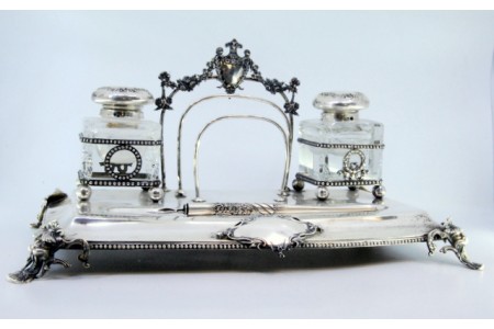 Tinteiro em prata com dois depósitos em vidro, caneta, suporte para cartas e assente sobre quatro pés de mascarões. 