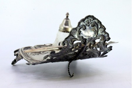 Tinteiro em prata recortada com um depósito em vidro, suporte para caneta e assente sobre três pés.