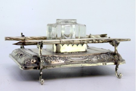 Tinteiro em prata relevado com motivos florais, friso perlado na envolvência, suporte para canetas, depósito em vidro e assente sobre quatro pés em forma de dragões.