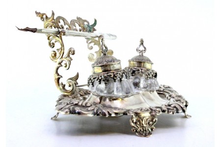 Tinteiro em prata relevada com dois depósitos em vidro, porta canetas e assente em quatro pés.