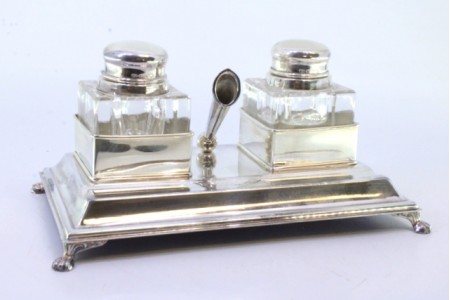 Tinteiro em prata liso com dois depósitos em vidro e suporte para caneta, assente em base lisa com friso e quatro pés em forma de garra com o fundo forrado.