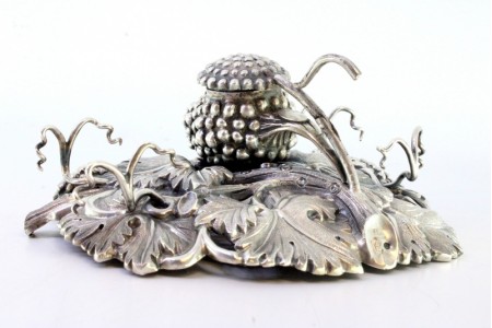 Tinteiro em prata com relevos em forma de cachos e paras, com dois suportes para canetas e tampa basculante.