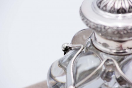 Tinteiro em vidro moldado com estrutura em prata, aplicações em meias bolas, tampa relevada com frisos e assente sobre base circular com cinco pés relevados.